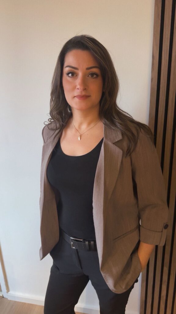 Manal Sultan står i ett professionellt porträtt vid en ljus vägg, klädd i kavaj och svart topp, med ett tryggt och seriöst uttryck.