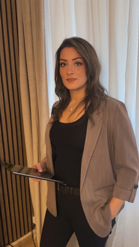 Manal Sultan stående med en iPad i handen framför ljusa gardiner, klädd i professionell outfit.